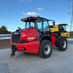 Front-left view of a red Manitou telehandler with large tires, yellow construction equipment behind, under a blue sky with power lines.
