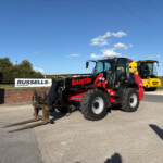 Red Manitou telehandler with long forks at a construction lot, Russell's sign in the background under a blue sky.