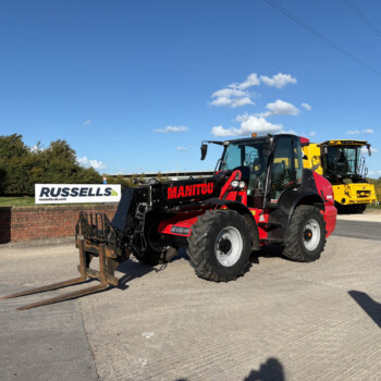 Red Manitou telehandler with long forks at a construction lot, Russell's sign in the background under a blue sky.