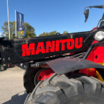 Close-up of a red and black Manitou crane with large tires and headlights against a clear blue sky, flags in background.