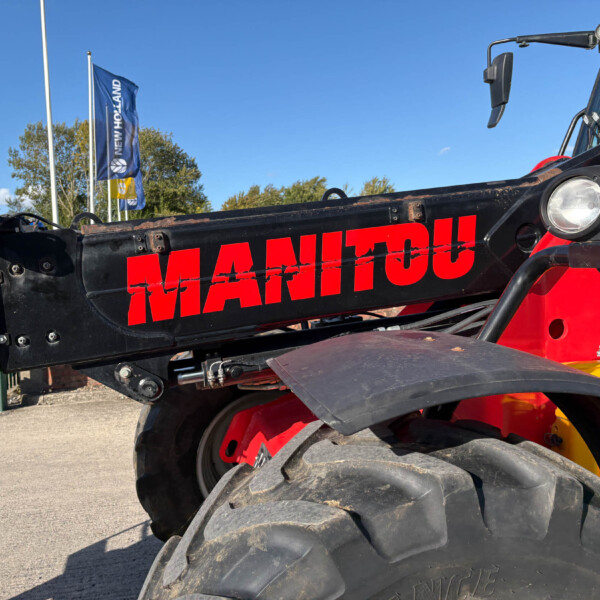 Close-up of a red and black Manitou crane with large tires and headlights against a clear blue sky, flags in background.