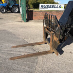 Forklift forks extended toward the viewer in a yard, with a Russell's sign and industrial equipment in the background.