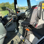 Interior of a Manitou construction vehicle cab with steering wheel, orange control lever, and dusty floor mats visible at the doorway.