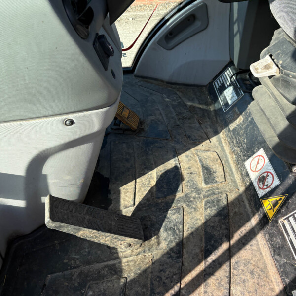 Footwell of a dusty construction vehicle cabin showing pedals and a worn floor mat with safety stickers nearby on the floorboard