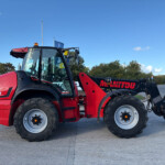 Red Manitou telehandler with large tires and lifted arm, parked on a paved lot under a blue sky.