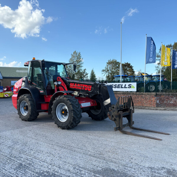 Red Manitou telehandler with forks attached, parked on a paved lot beside a brick wall and industrial buildings under a blue sky with clouds.