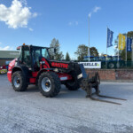 Red New Holland tractor with long forklift forks in a paved lot, blue sky above, and flags in the background to the right.