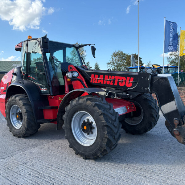 Red Manitou telehandler with bucket attachment parked in an outdoor yard against a blue sky with flags in the background; construction equipment area visible.