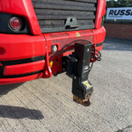 Close-up of the red truck nose with a black grille, round headlights, and a hanging tow hitch in the foreground; a Russell sign is visible in the background.