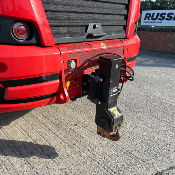 Close-up of the red truck nose with a black grille, round headlights, and a hanging tow hitch in the foreground; a Russell sign is visible in the background.