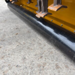 Close-up of a yellow snow removal attachment with orange rusted paddles and a black curved cutting edge resting on concrete