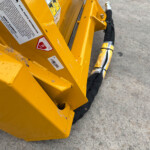 Close-up of yellow construction machinery frame with safety stickers and a black-wrapped hose on a concrete surface.