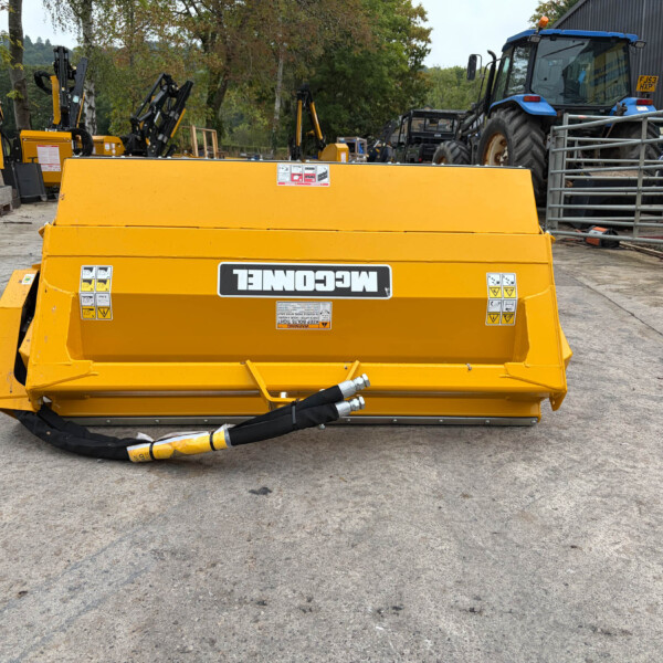 Front view of a large yellow agricultural/industrial mower attachment in a yard with multiple machines and a blue tractor in the background.