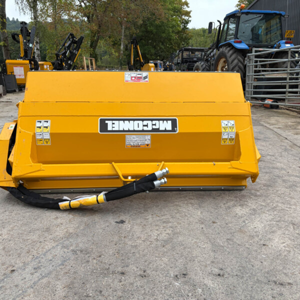 Front view of a large yellow construction compactor in a yard, with other machinery and a blue tractor in the background.