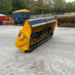 Yellow agricultural header with rotating blades and rollers, parked in an outdoor equipment yard with other machines in the background.