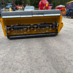 Yellow front-mounted industrial attachment with a black roller and metal tines, situated in a yard with various tractors and heavy equipment in the background