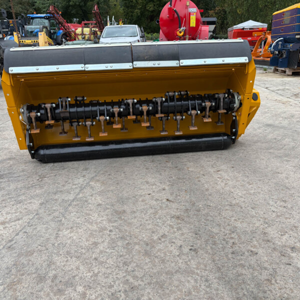 Yellow front-mounted industrial attachment with a black roller and metal tines, situated in a yard with various tractors and heavy equipment in the background