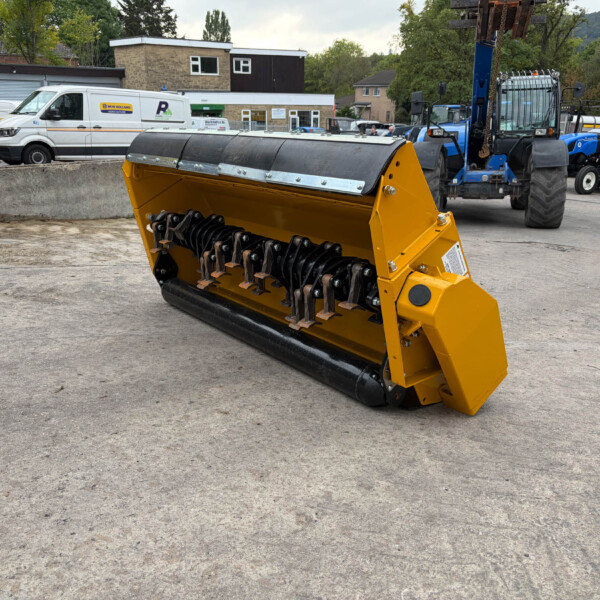 Yellow industrial snow blower attachment in the foreground with machinery and vans in a yard behind it, outdoor construction site setting.