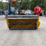 Large yellow street-sweeping machine foreground with bristles, parked on a concrete lot; background shows blue tractors, trailers, and a red agricultural unit amid trees.