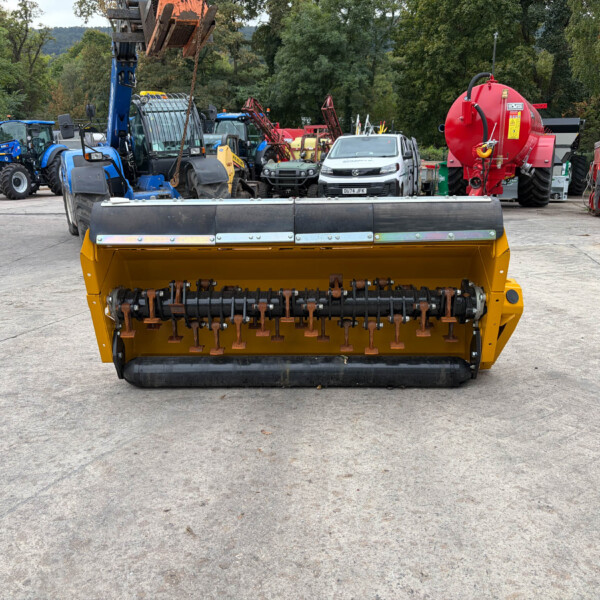 Large yellow street-sweeping machine foreground with bristles, parked on a concrete lot; background shows blue tractors, trailers, and a red agricultural unit amid trees.