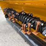 Close-up of a yellow industrial machine with rusty black/silver roller gears and mounting bolts along the base rail. Need for maintenance evident by rusted components. Alternative: A yellow machine frame exposing rusted roller teeth and linkages along a conveyer-like assembly.