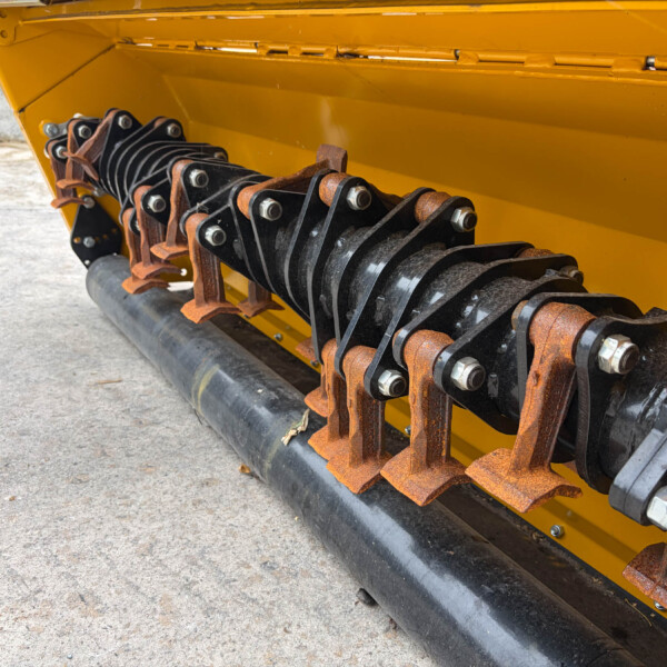 Close-up of a yellow industrial machine with rusty black/silver roller gears and mounting bolts along the base rail. Need for maintenance evident by rusted components. Alternative: A yellow machine frame exposing rusted roller teeth and linkages along a conveyer-like assembly.