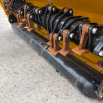 Close-up of rusted metal rollers and belts on a yellow industrial machine's undercarriage.