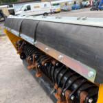 Close-up of a yellow construction machine’s roller system with metal guards and bolts; rusted gears visible beneath.