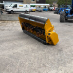 Yellow industrial snow blower attachment on a concrete surface with parked vans and blue heavy machinery in a yard behind it.