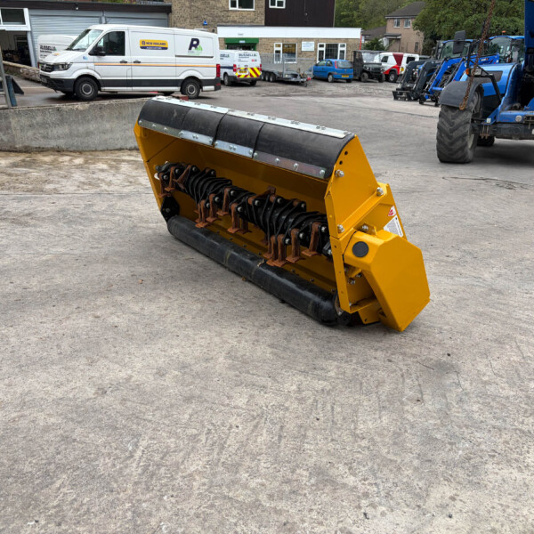 Yellow industrial snow blower attachment on a concrete surface with parked vans and blue heavy machinery in a yard behind it.