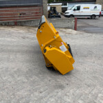 Close-up of a yellow industrial attachment on wheels in a gravel yard, with white vans in the background.
