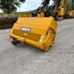 Yellow industrial mulching/cutting attachment on a concrete yard, with other yellow machines and a rural field in the background.