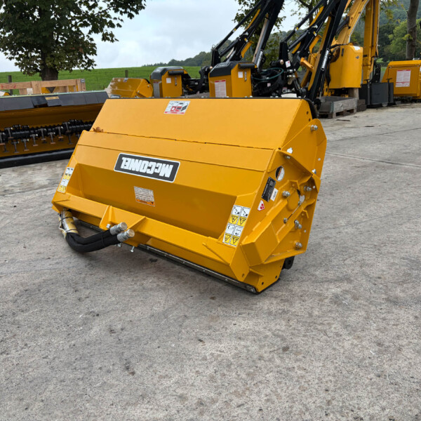Yellow industrial mulching/cutting attachment on a concrete yard, with other yellow machines and a rural field in the background.