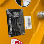 Close-up of a yellow industrial machine showing a black data plate with a QR code, serial numbers, and a red '8 hrs' warning sticker nearby.