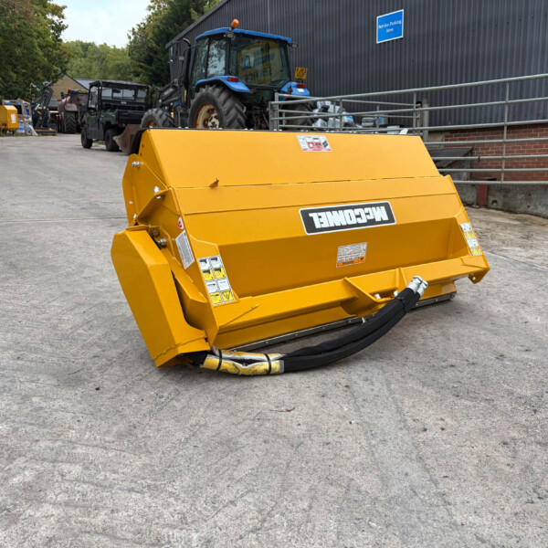 Bright yellow heavy-duty agricultural attachment with hydraulic hoses on a concrete yard; tractors and equipment line the background near a dark metal building.