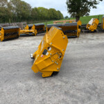 Row of yellow industrial machinery attachments parked on a concrete lot with trees in the background.