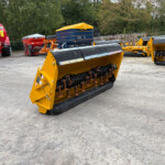 Yellow agricultural implement with metal rollers in foreground at an outdoor equipment yard, with red tank and other machinery in the background
