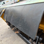 Close-up of a dark metal panel on a yellow industrial machine, showing scuffs and scratches, with a blurred outdoor background and vehicles.