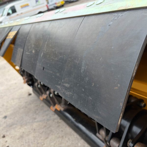 Close-up of a dark metal panel on a yellow industrial machine, showing scuffs and scratches, with a blurred outdoor background and vehicles.