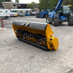 Yellow industrial snow blower attachment in the foreground with a yard of construction machinery and vans in the background.