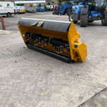 Yellow industrial snow plow attachment on a concrete yard with blue tractors and service vans in the background