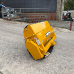 Tilted yellow industrial waste hopper on wheels in a construction yard, with a blue tractor and metal railing nearby and a brick building in the background.