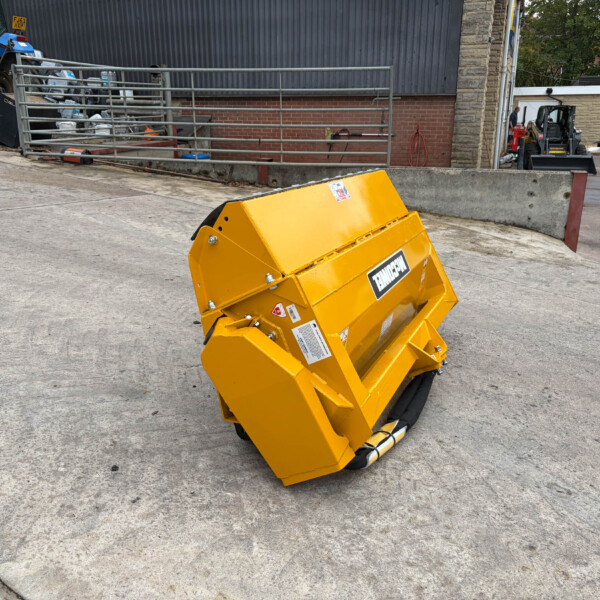 Tilted yellow industrial waste hopper on wheels in a construction yard, with a blue tractor and metal railing nearby and a brick building in the background.