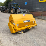 Yellow agricultural attachment in the foreground, with blue tractors and a service building in the background at an outdoor equipment yard.