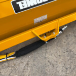 Close-up of yellow construction equipment with warning labels and hydraulic hoses on a concrete surface.