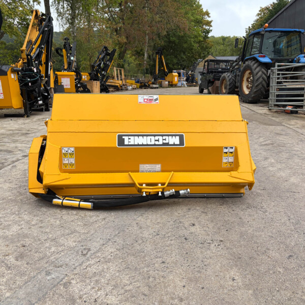 Front view of a large yellow agricultural machine in a yard, with other yellow and black attachments and a blue tractor in the background.