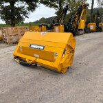 Row of yellow construction machines with hydraulic arms parked on a paved surface, with trees and fields in the background.