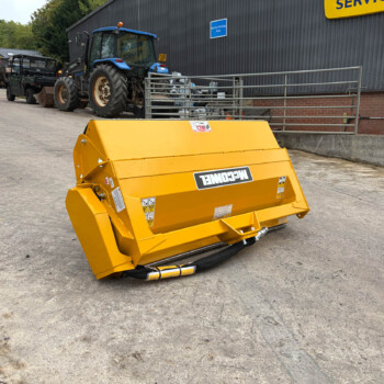 Yellow agricultural attachment in the foreground, with blue tractors and a service building in the background at an outdoor equipment yard.