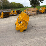 Row of bright yellow agricultural or industrial machine attachments lined up on a gravel lot, with pallets and a grassy, tree-lined field in the background.