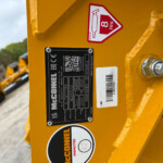 Close-up of yellow construction equipment showing safety and identification stickers on the side, including a black plate and a red warning sign nearby.
